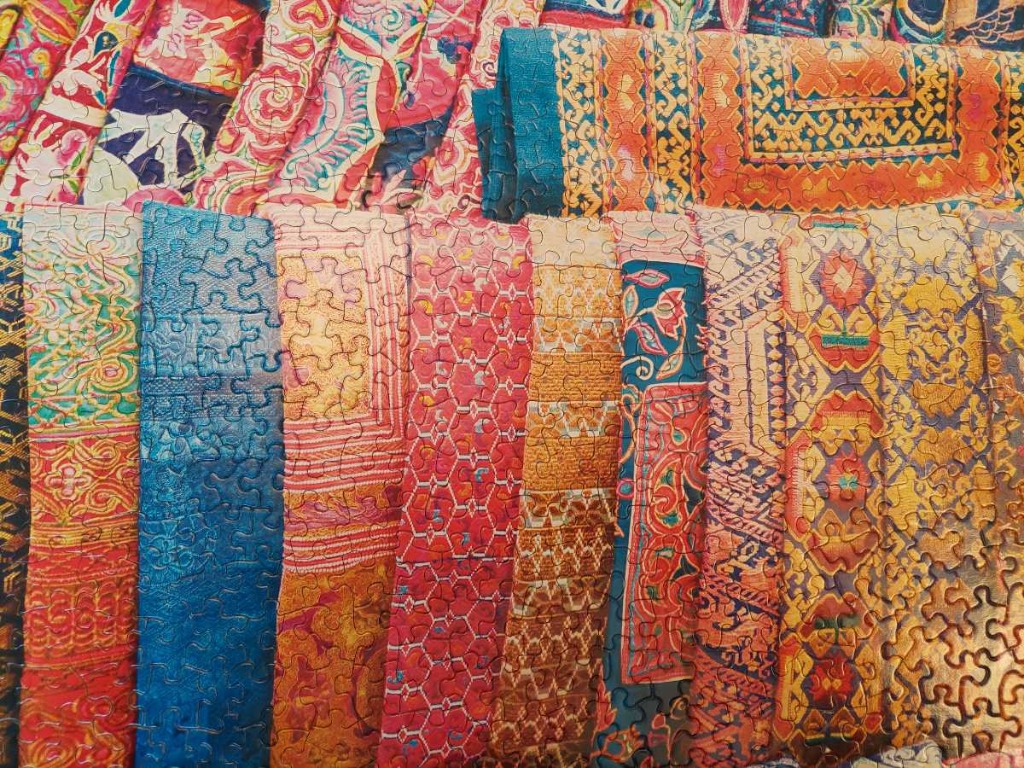 A vibrant collage of folded textile fabrics featuring ornate, traditional patterns in warm reds, oranges, blues, and golds.
