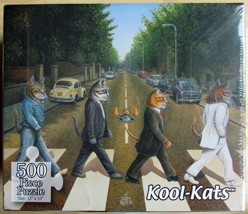 Four cats dressed in suits and casual clothing walking across a crosswalk on a tree-lined city street.