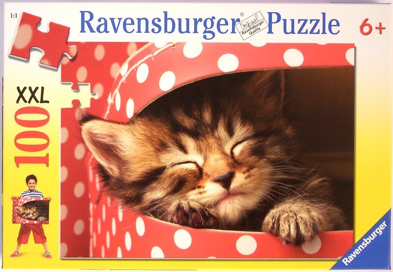 A close-up photograph of a fluffy tabby kitten sleeping inside a bright red polka-dotted box.