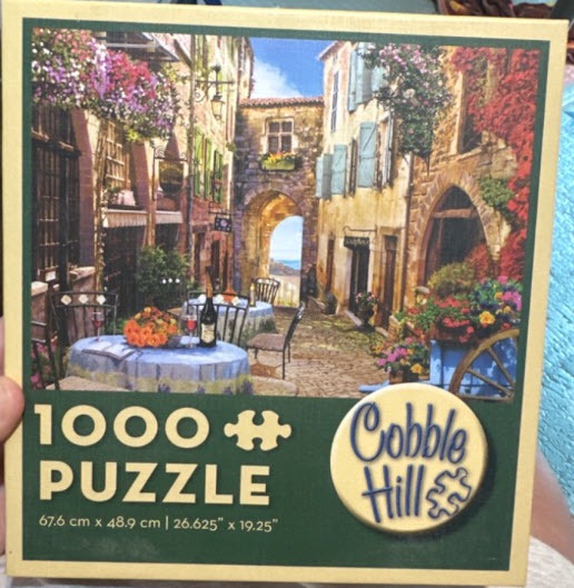 A scenic painting of a quaint European cobblestone alleyway with outdoor café seating and vibrant flower displays.