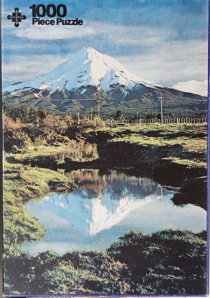 Photograph of Mount Taranaki reflected in a still pool in a grassy field with a fence in the mid-ground.