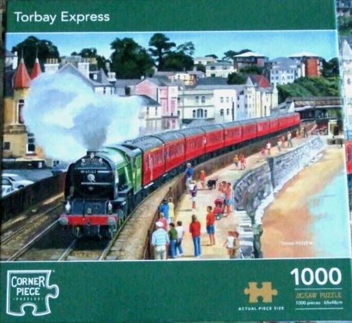 A steam train pulling red passenger cars along a coastal promenade with buildings in the background.