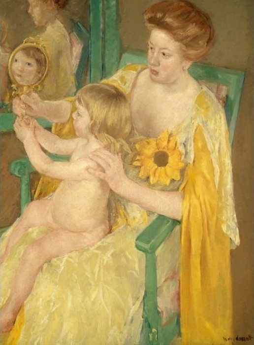 A woman in a yellow dress holding a small toddler who looks into a hand mirror.