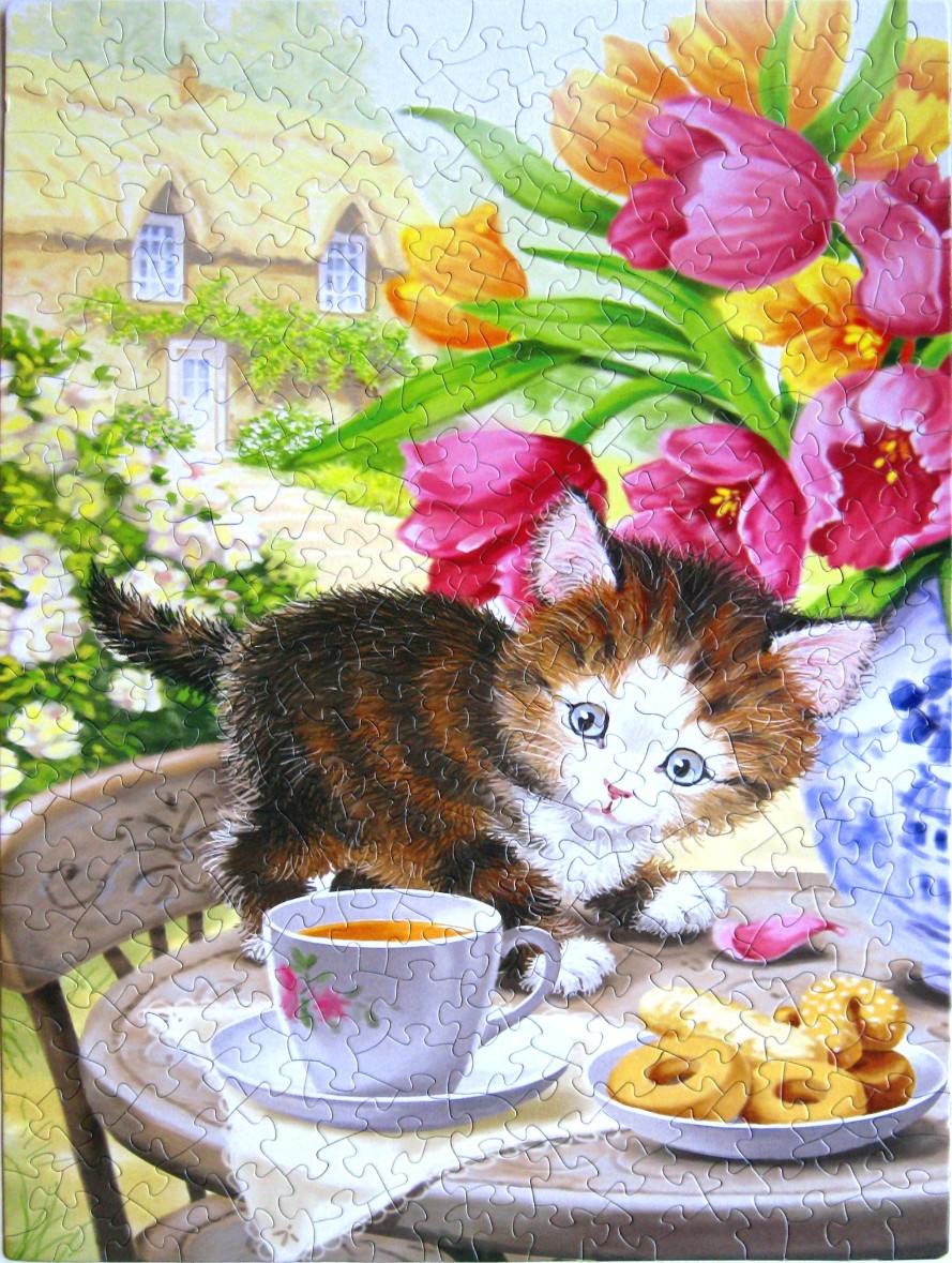 A fluffy kitten sits on a garden table next to a cup of tea, flowers, and cookies with a cottage in the background.