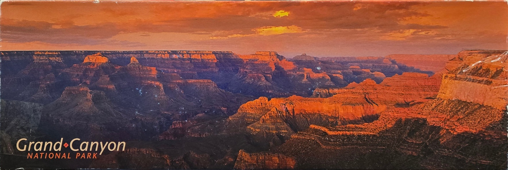 A panoramic landscape photograph of the Grand Canyon at sunset with warm orange, red, and deep blue shadows.