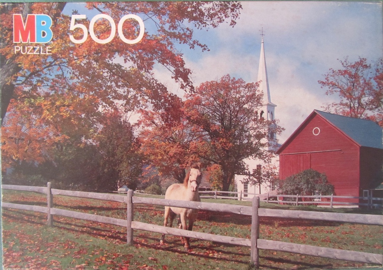 A light-colored pony stands in a fenced pasture in front of a red barn and white church steeple during autumn.