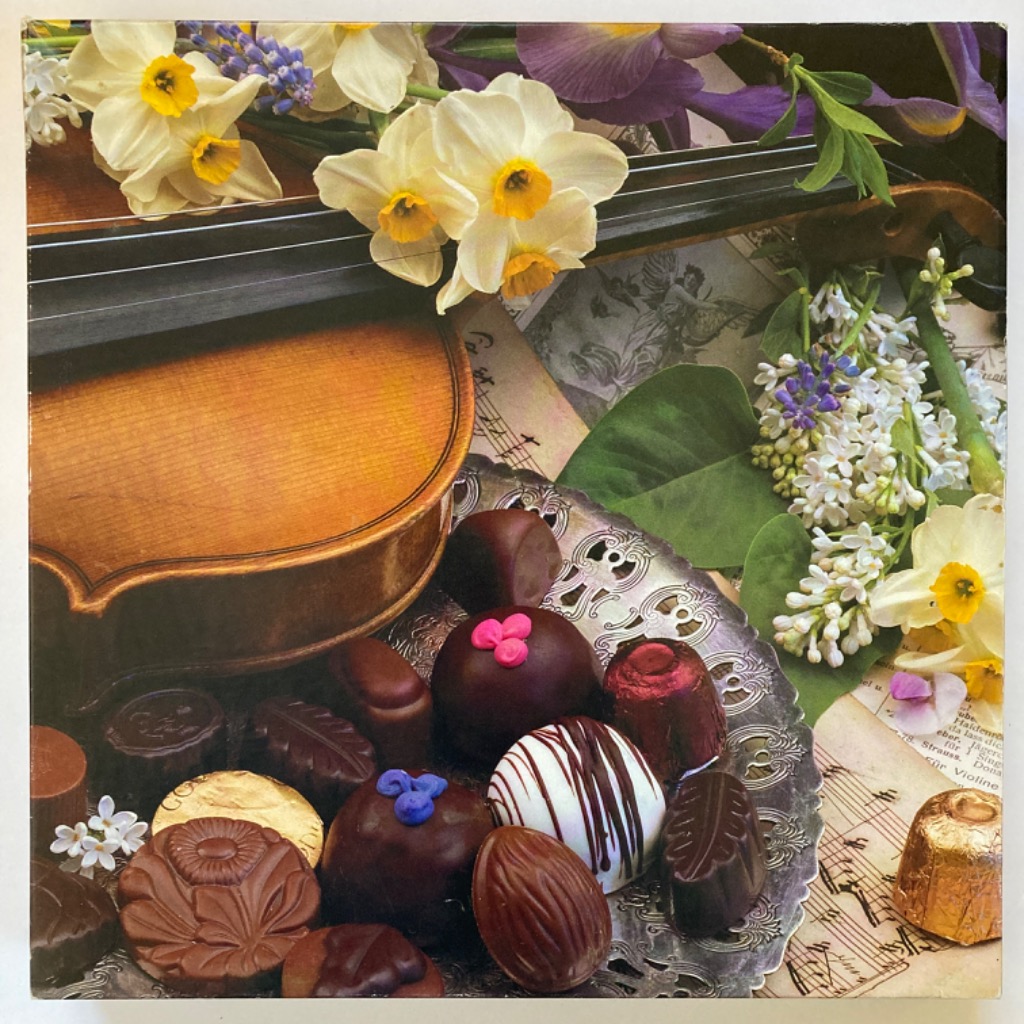 A still life featuring a wooden violin, assorted chocolates on a silver platter, and spring flowers.