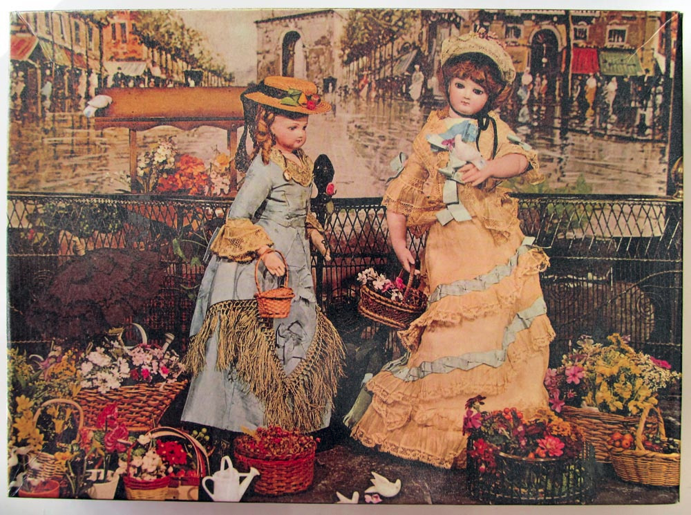 Two elegant Victorian-style dolls in fancy dresses standing in a flower market with a city street background.