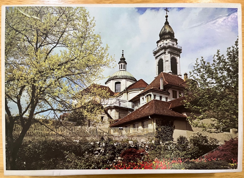 A scenic view of a baroque-style church with a prominent dome and tall bell tower, framed by trees and a garden.