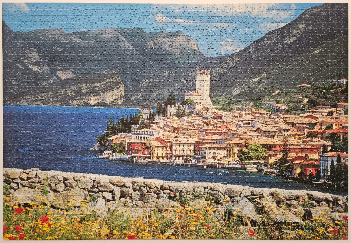 A Mediterranean lakeside town with a prominent castle tower, backed by mountains, with wildflowers in the foreground.