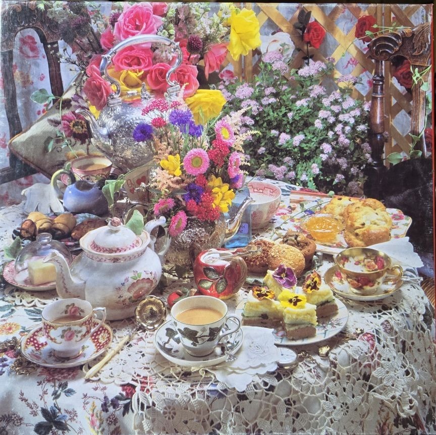 A lush garden tea party scene featuring silver teapots, floral arrangements, pastries, and fine china.