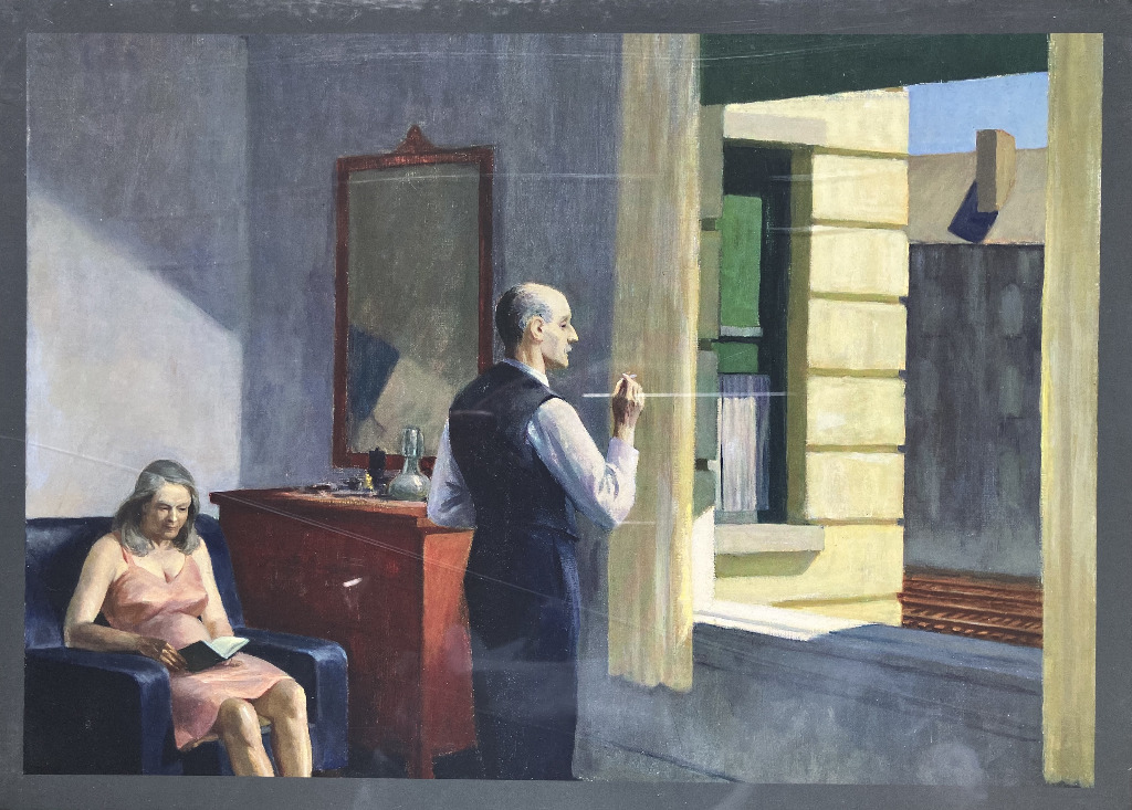 A woman reads in an armchair while a man stands by a window in a dimly lit, somber room.
