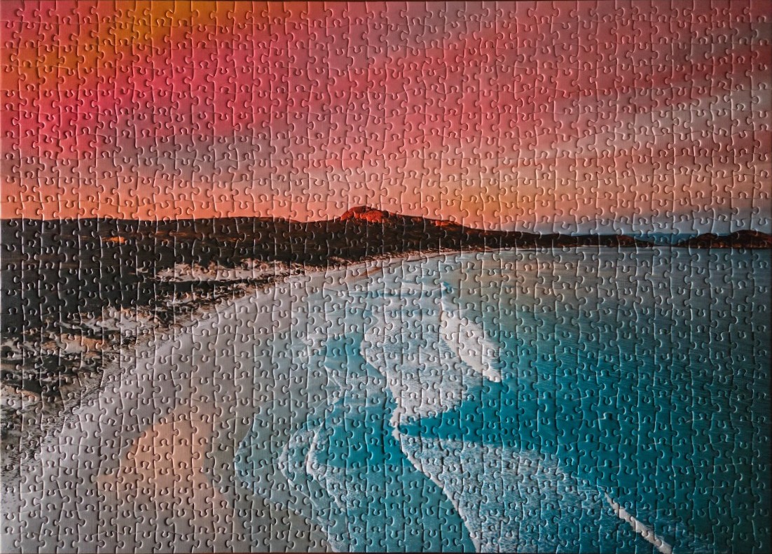 An aerial view of a coastline at sunset, with turquoise water meeting a sandy beach and hills under a pink sky.