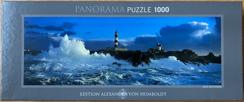 A panoramic photograph of a lighthouse surrounded by a turbulent, crashing sea under a deep blue twilight sky.