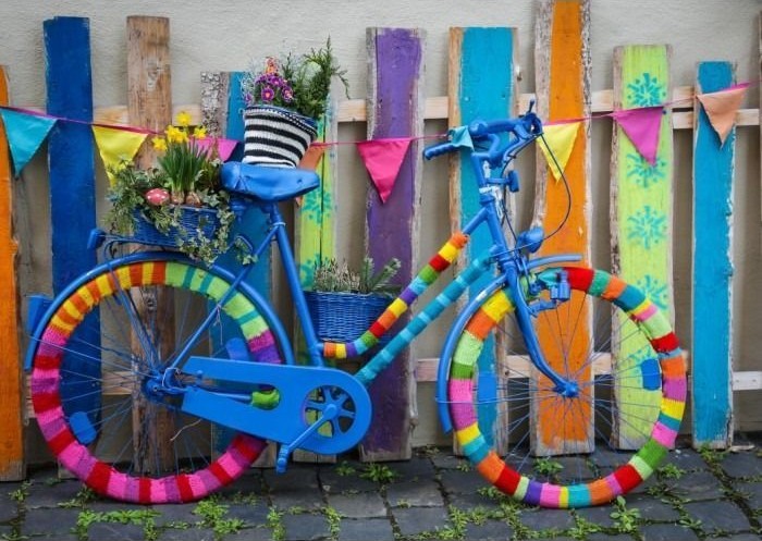 A blue bicycle decorated with colorful knitted yarn and flower baskets parked against a vibrant, multi-colored wooden fence.