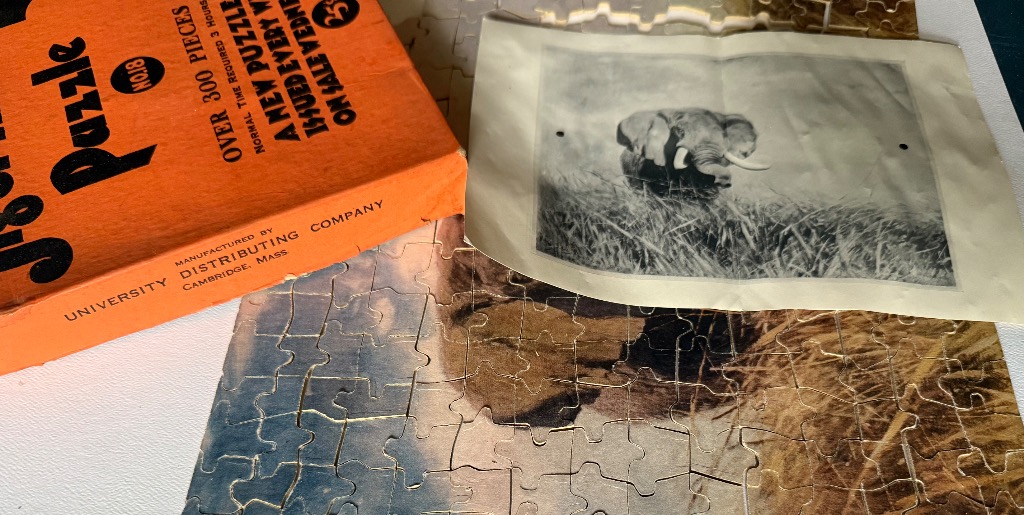 An orange box and a paper photograph of an elephant resting on an incomplete surface of puzzle pieces.