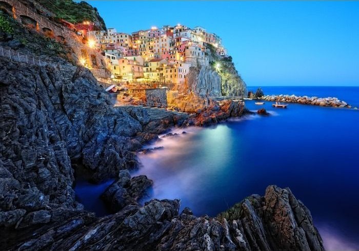 A cliffside Italian coastal village at dusk with glowing lights illuminating the buildings above the dark sea.