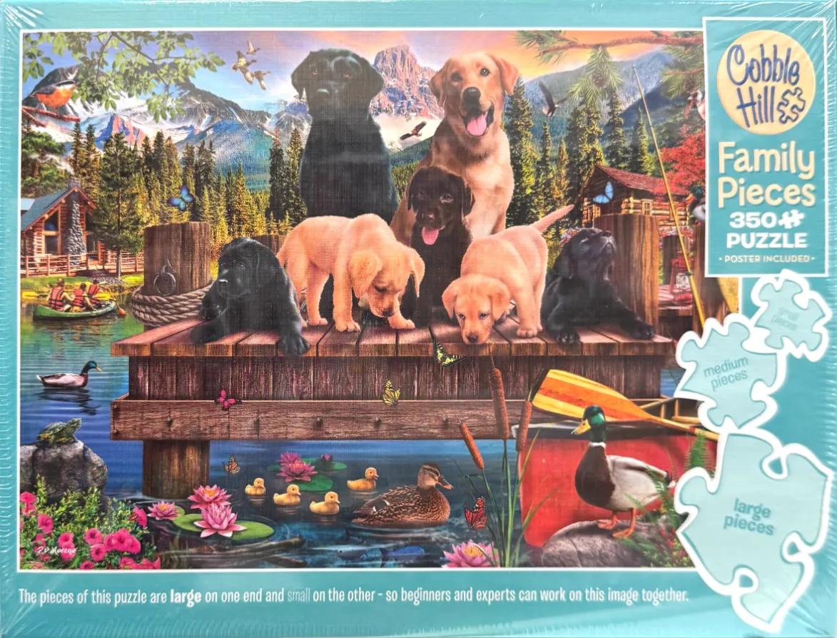 Seven Labrador puppies and adult dogs sitting on a wooden dock by a lake with a mountain backdrop.