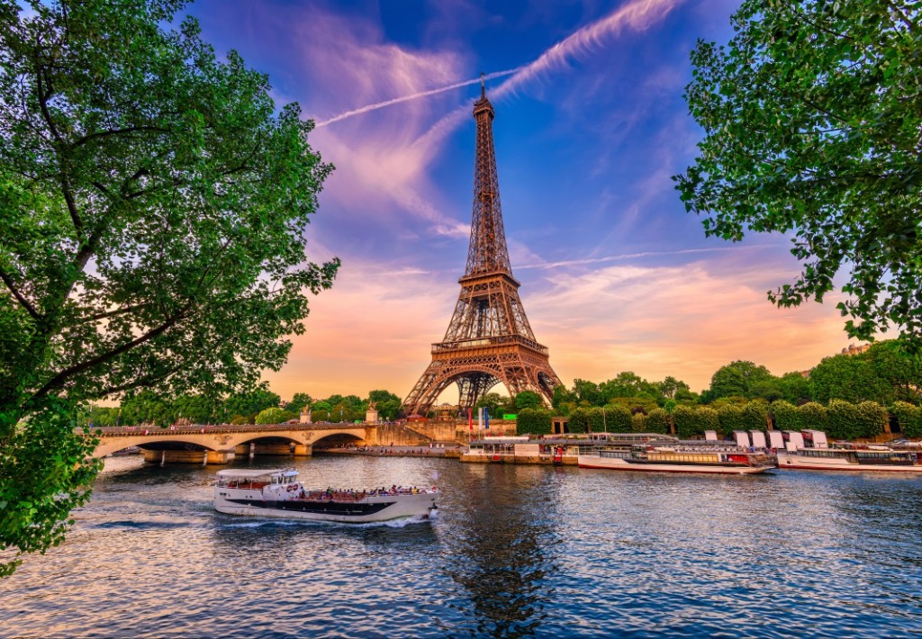 The Eiffel Tower rises above the Seine River at sunset, framed by lush trees and riverboats.
