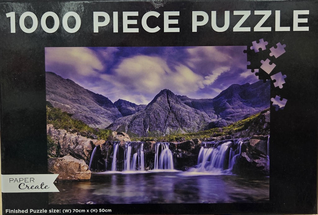 A panoramic photograph of a cascading waterfall flowing beneath jagged, purple-toned mountains under a dramatic sky.