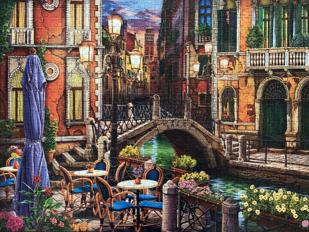 A charming Venetian canal-side cafe at twilight, featuring iron lamps, stone bridges, and historic orange buildings.
