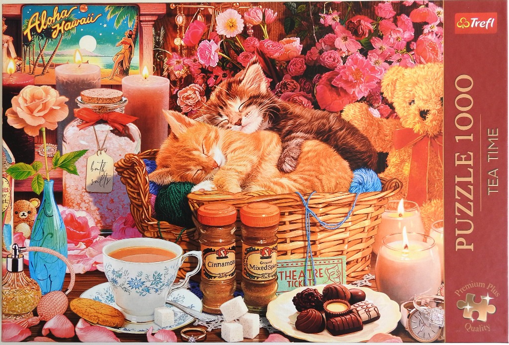 Two kittens sleeping in a wicker basket surrounded by tea, sweets, flowers, and candles.
