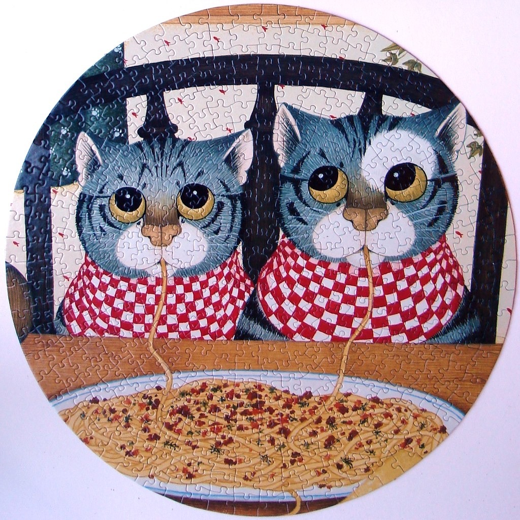 Two cartoon cats wearing red and white checkered scarves eating spaghetti from the same plate.