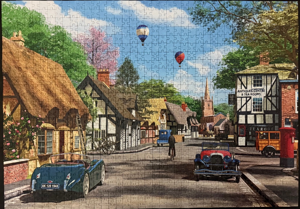 A nostalgic painting of an English village street featuring antique cars, thatched cottages, and hot air balloons.