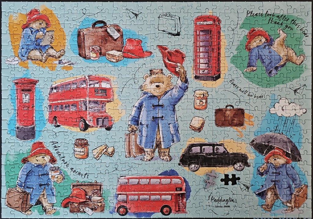 A whimsical collage of Paddington Bear, red London buses, phone boxes, and suitcases on a light blue background.