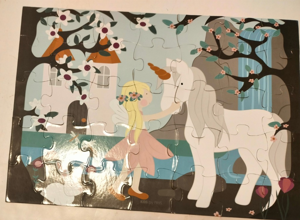 A whimsical illustration of a blonde girl with wings interacting with a white unicorn in front of a castle and waterfall.