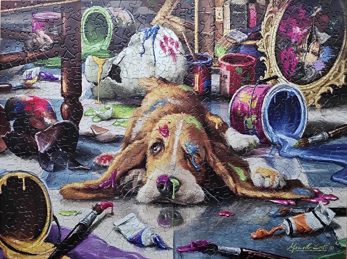 A Basset Hound puppy covered in multi-colored paint sits amidst a chaotic studio floor full of spilled art supplies.