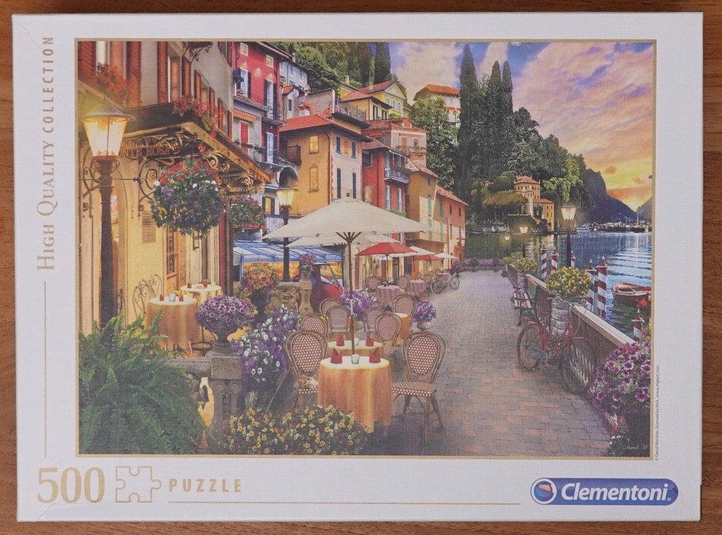 A charming Mediterranean-style lakeside terrace with dining tables, colorful buildings, and a sunset over the water.
