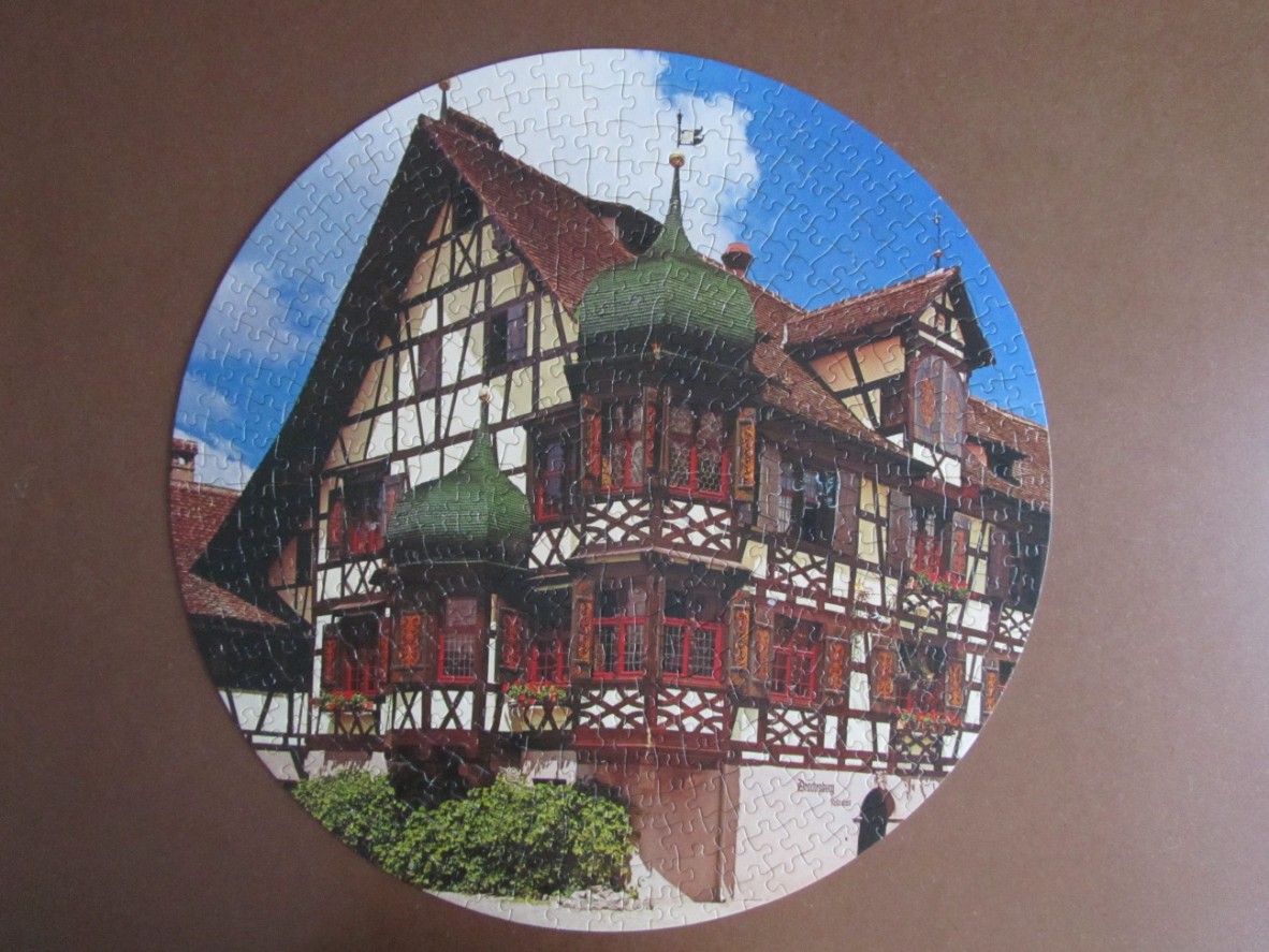 A circular image of a traditional half-timbered German house with two ornate green turrets against a blue sky.