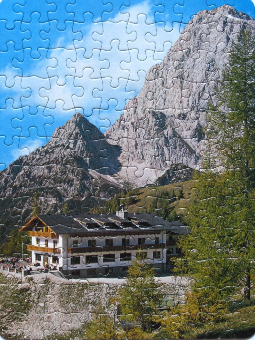 A traditional mountain hotel nestled at the base of dramatic, rocky alpine peaks under a bright blue sky.
