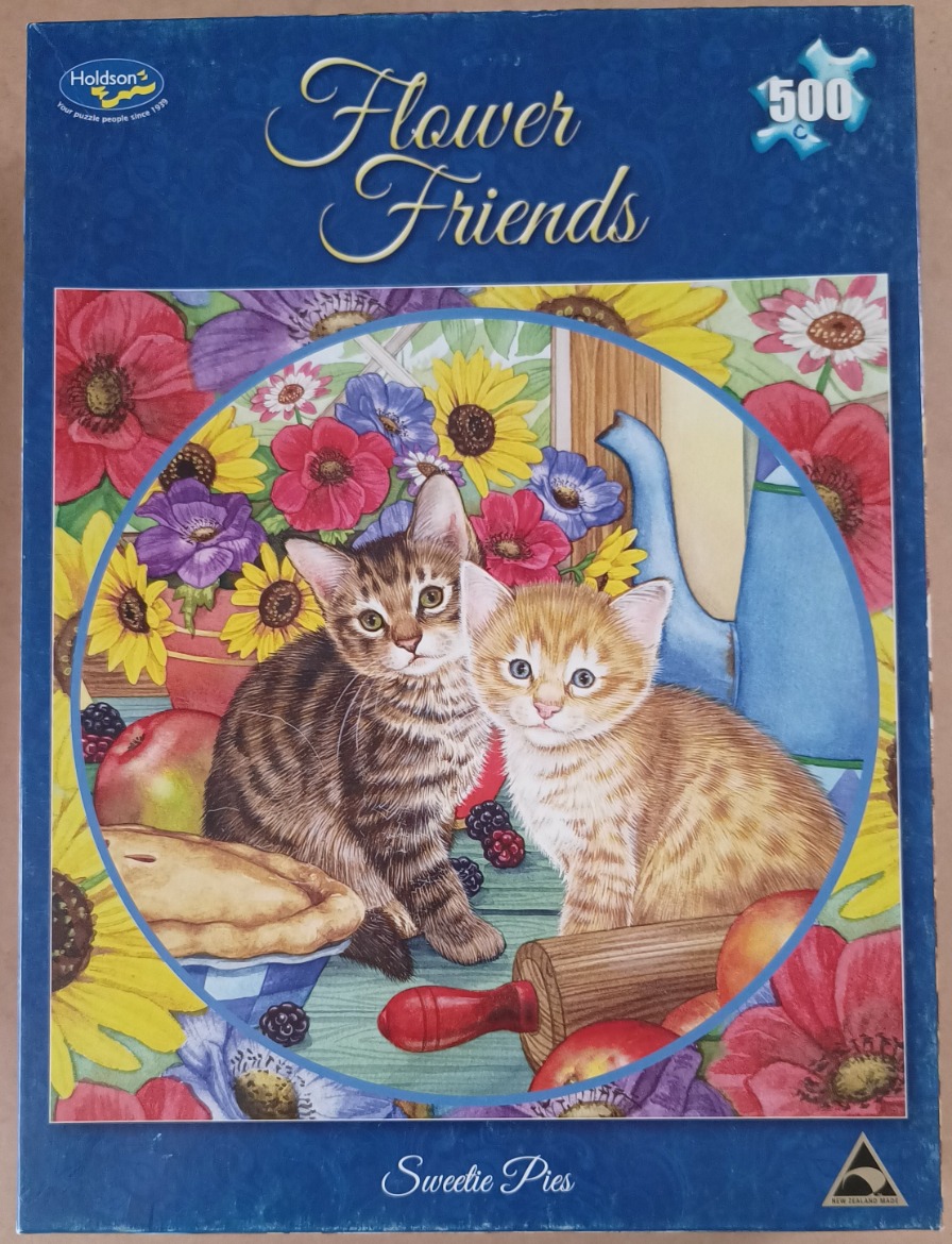 A tabby kitten and an orange tabby kitten sit on a wooden table surrounded by flowers, fruit, and a pie.