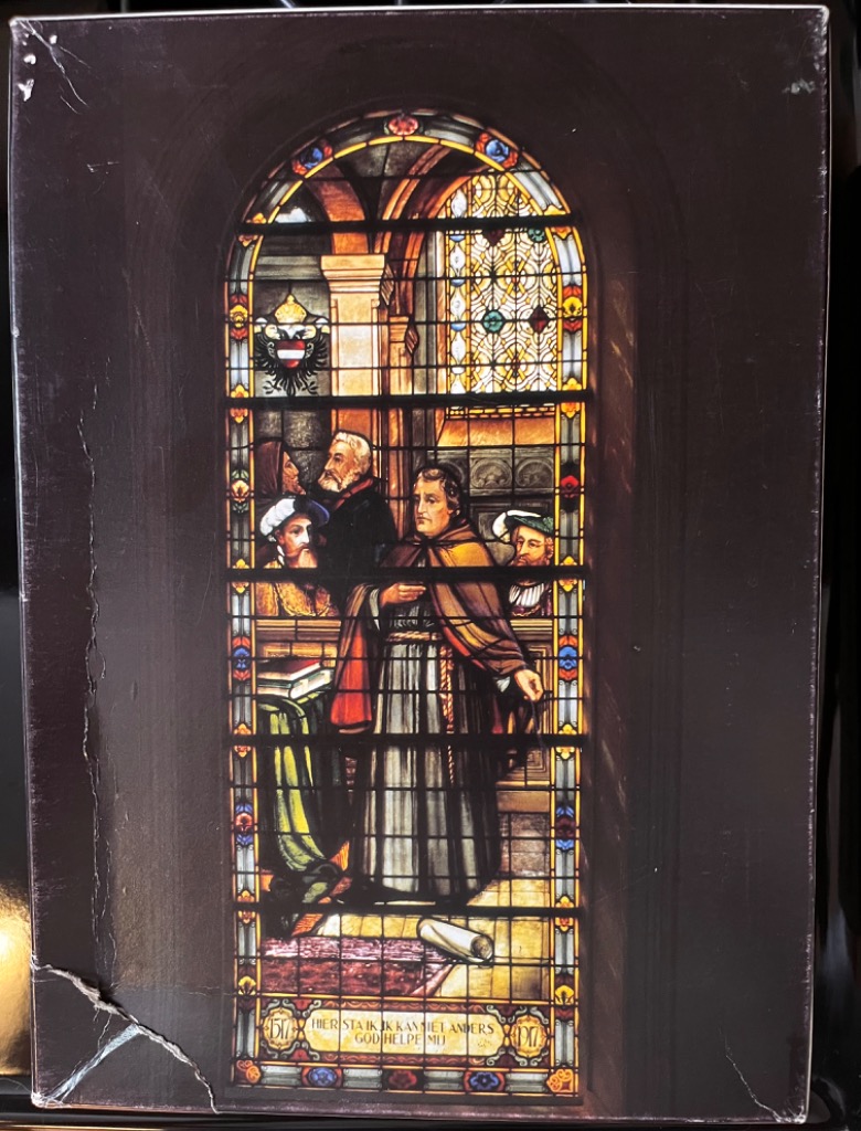 Stained glass artwork depicting Martin Luther standing before an audience, set against a dark background.