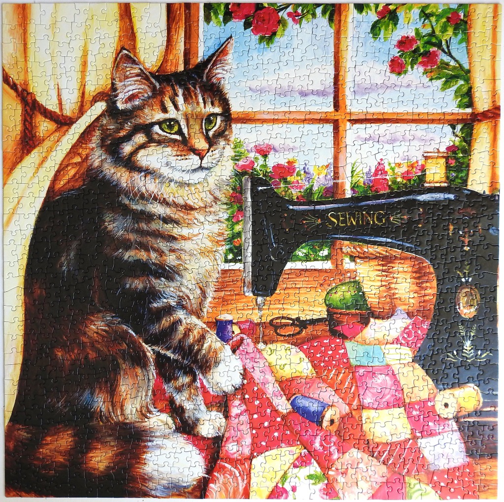 A fluffy tabby cat sits next to an antique black sewing machine and a colorful patchwork quilt by a sunny window.