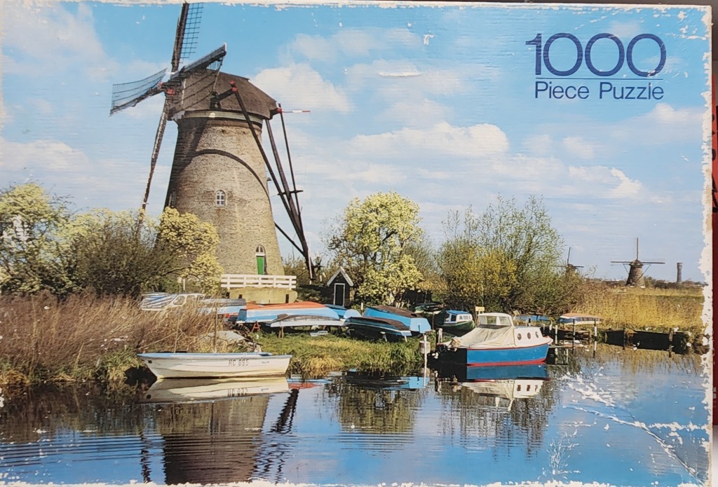 A traditional stone windmill overlooking a riverbank with small boats on a sunny day.