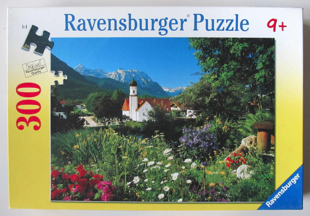 A sunny, scenic photograph of a white church with a tall steeple, surrounded by a lush garden and jagged alpine mountains.