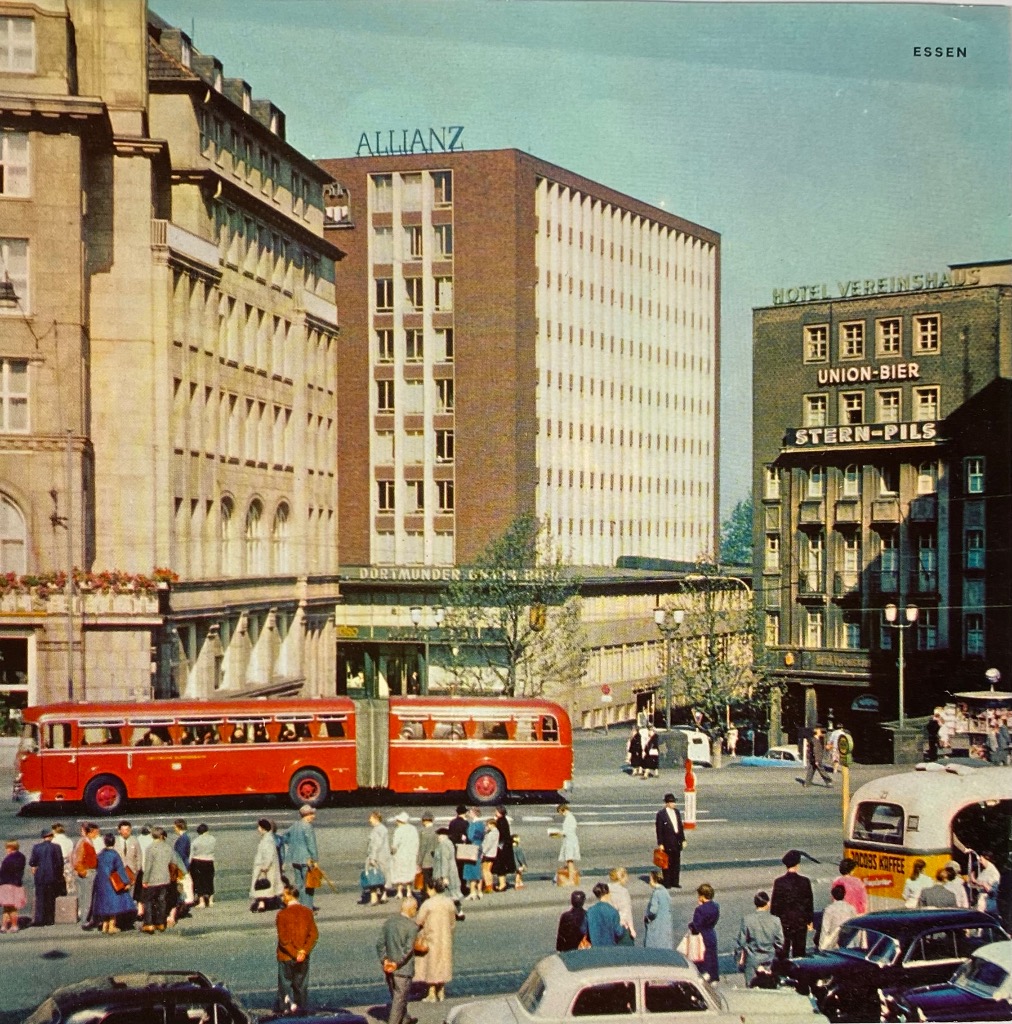 A street scene in Essen, Germany, featuring a long red articulated bus driving through a busy city intersection.