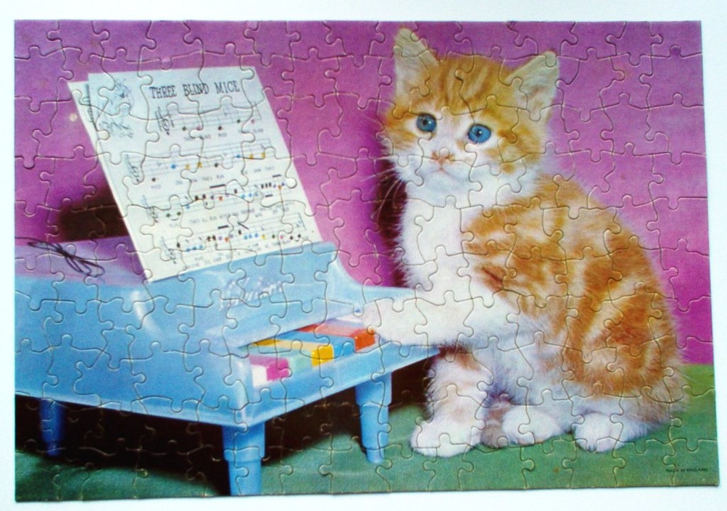 A ginger and white kitten sitting next to a light blue toy piano with sheet music for Three Blind Mice.