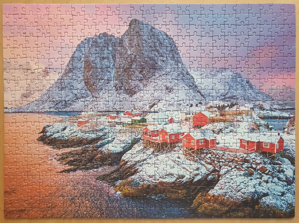 A coastal village with red cabins at the base of a snow-capped mountain under a pink and purple twilight sky.