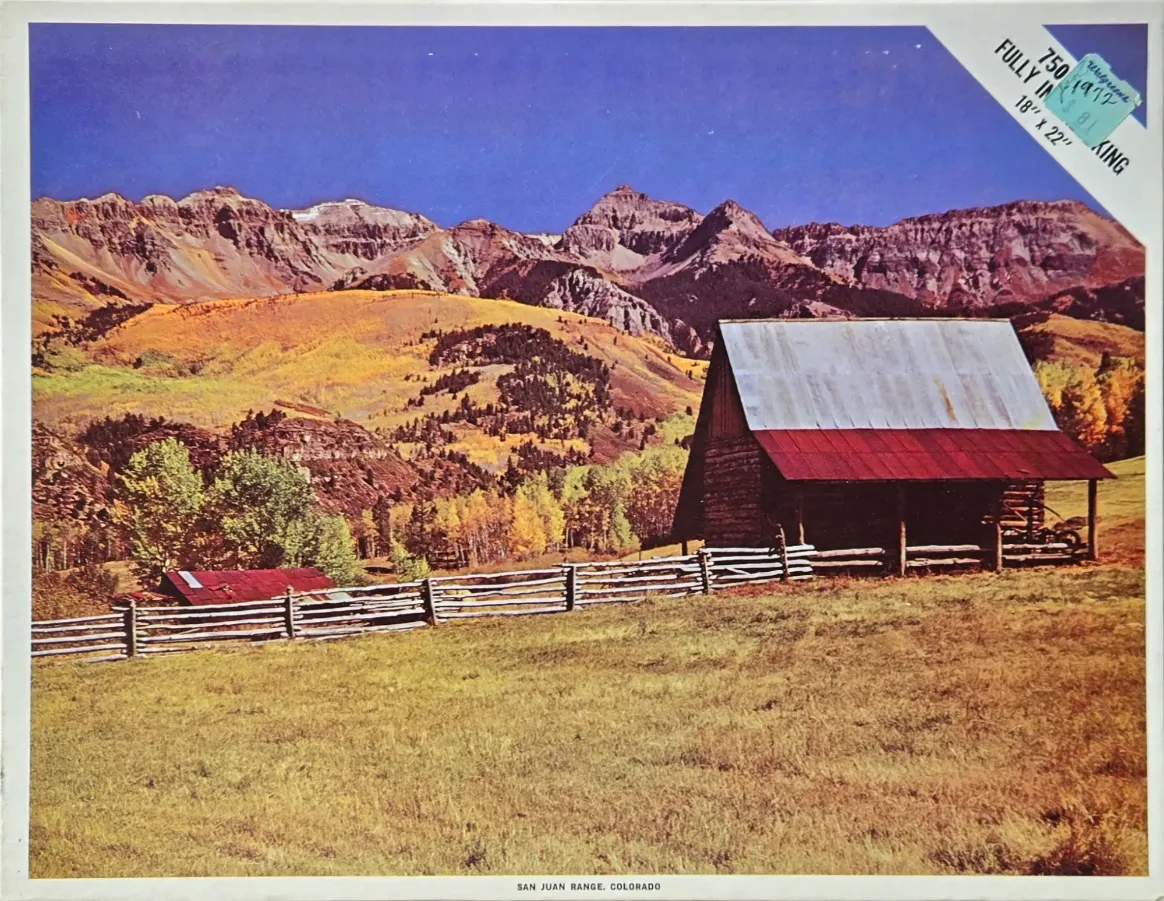A rustic wooden barn sits in a golden mountain meadow beneath the jagged peaks of the San Juan Range in Colorado.