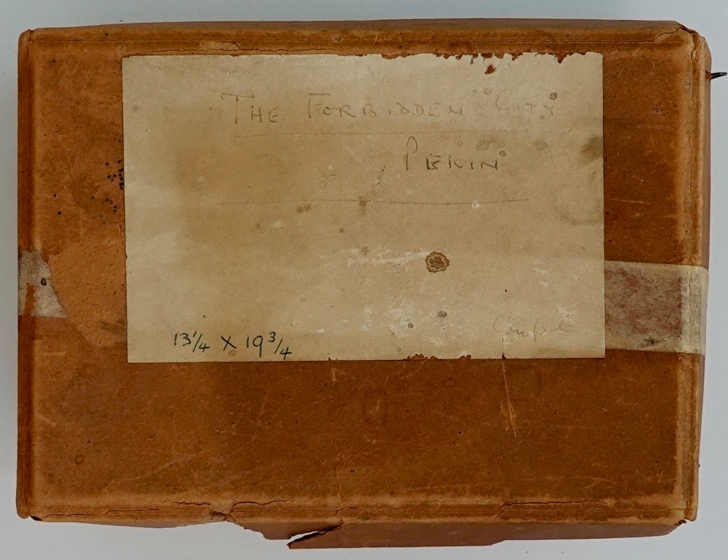 A rectangular, worn brown container with a handwritten paper label reading The Forbidden City, Pekin.