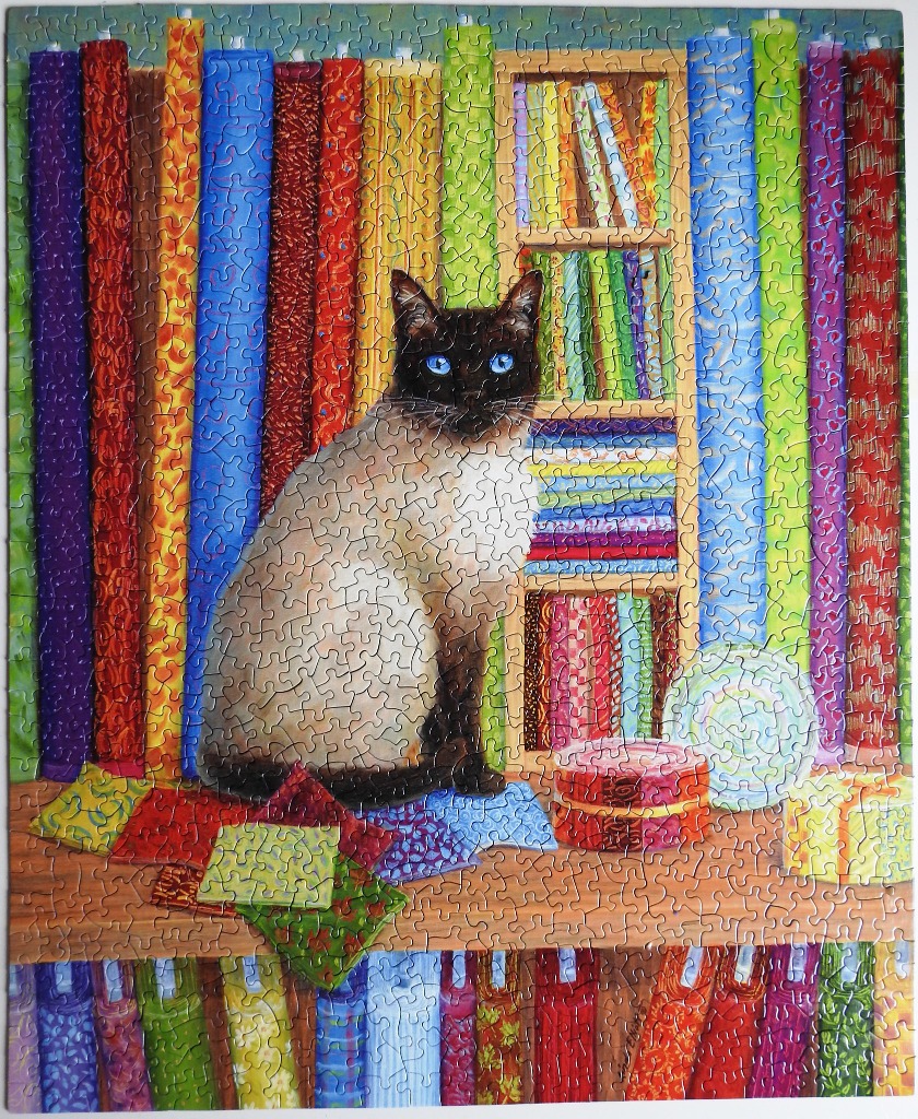 A Siamese cat sitting in a colorful craft room surrounded by fabric rolls and sewing supplies.