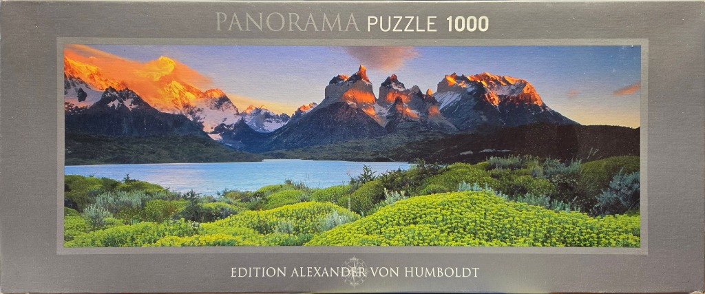 A panoramic photograph of the granite peaks of Torres del Paine glowing at sunrise above a blue lake and green vegetation.