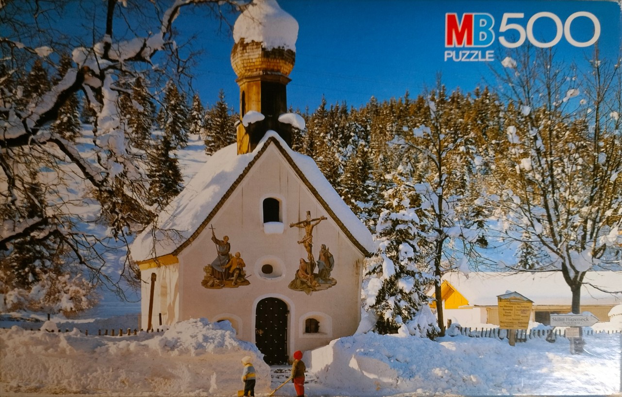 A small, snow-covered chapel in an alpine setting with two children clearing snow in front of the building.