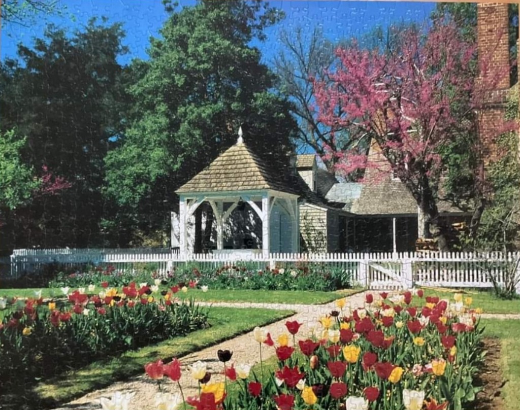 A sunny garden scene featuring a white gazebo, a rustic house, and flower beds filled with colorful tulips.