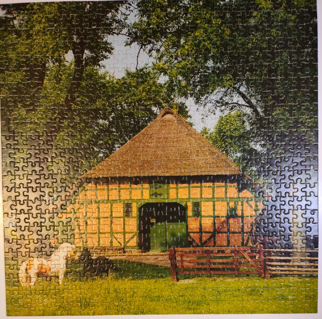 A timber-framed barn with a thatched roof stands in a sunny, green meadow under lush trees.