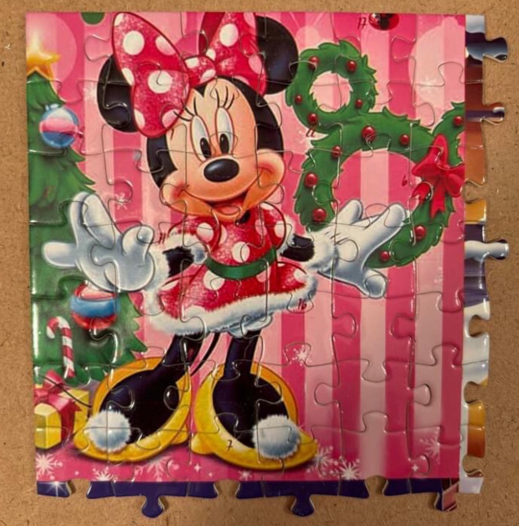 Minnie Mouse in a holiday dress holding a Mickey-shaped wreath with a Christmas tree in the background.